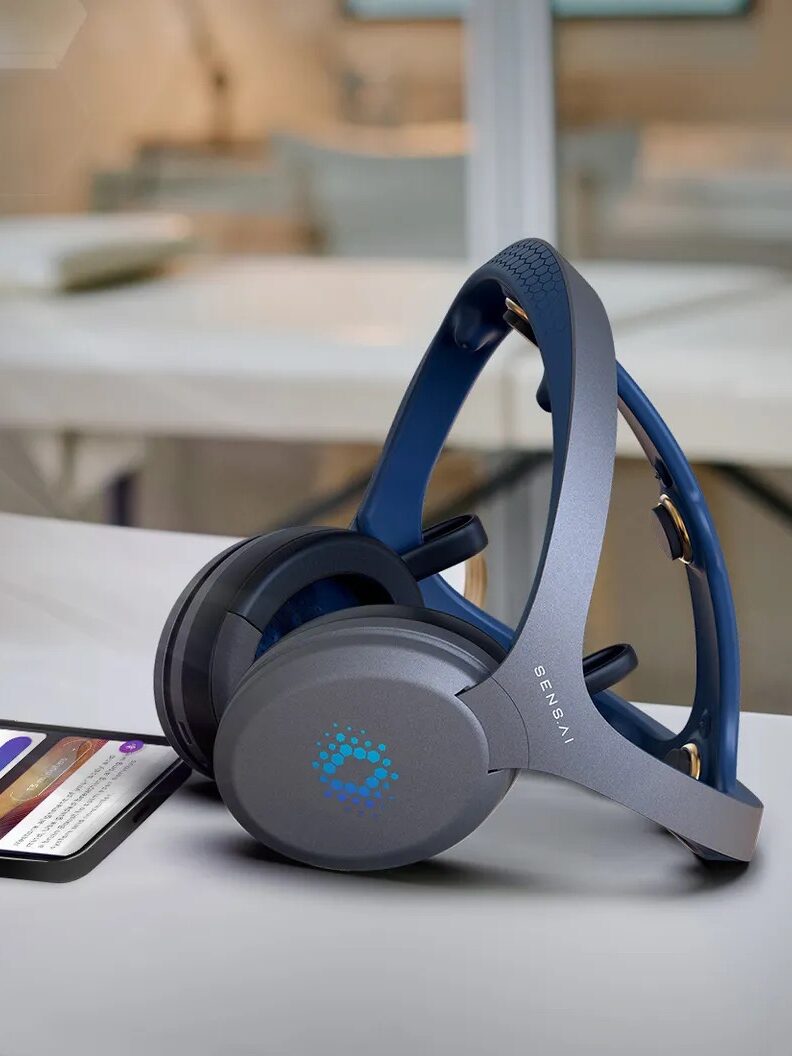 A pair of blue headphones rests on a white table next to a smartphone displaying a colorful app screen, with a blurred office background.