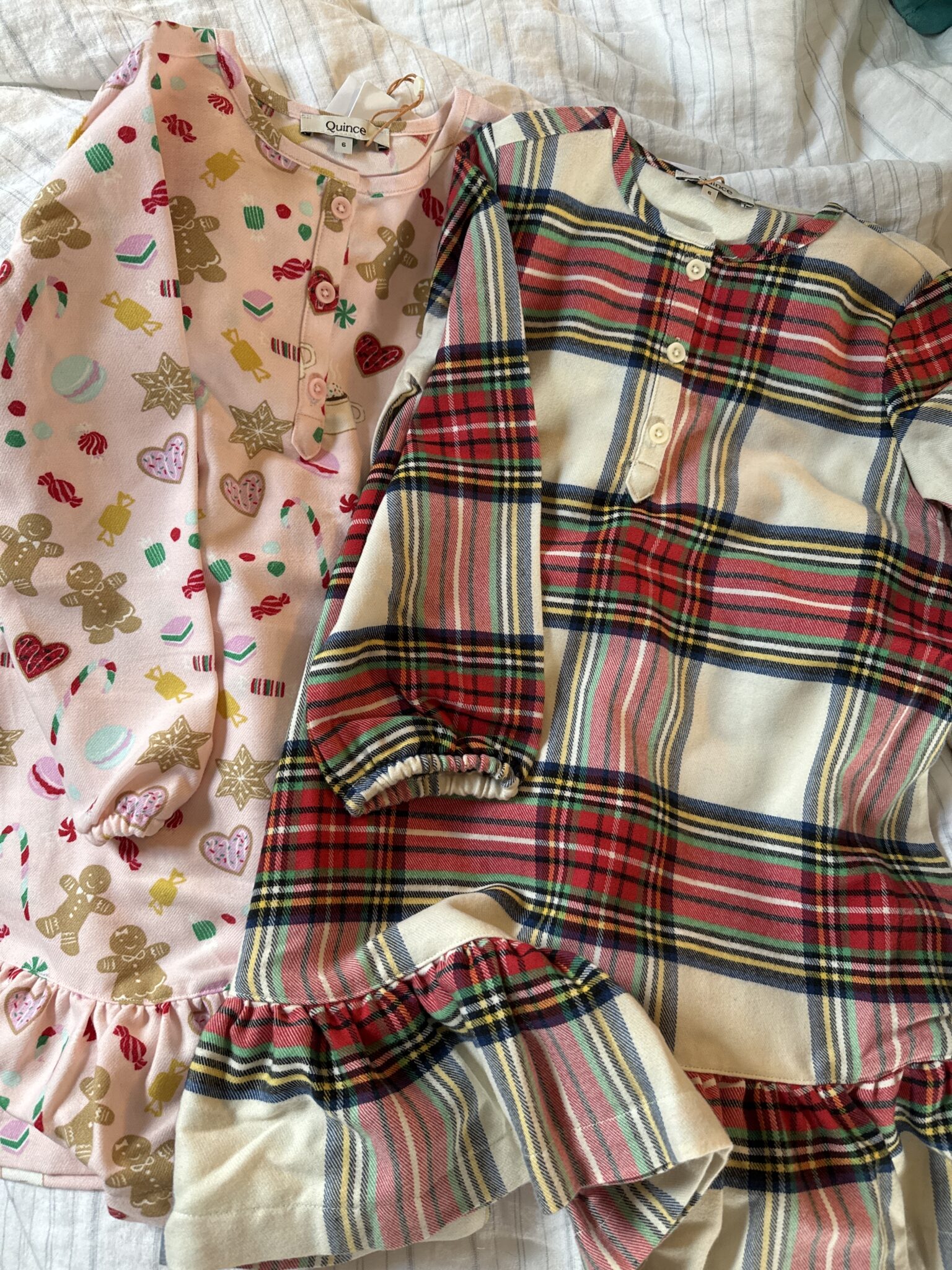 Two long-sleeve children's dresses lay side by side; one is pink with holiday cookie patterns, the other has a red, cream, and navy plaid design. Both have buttons on the front.