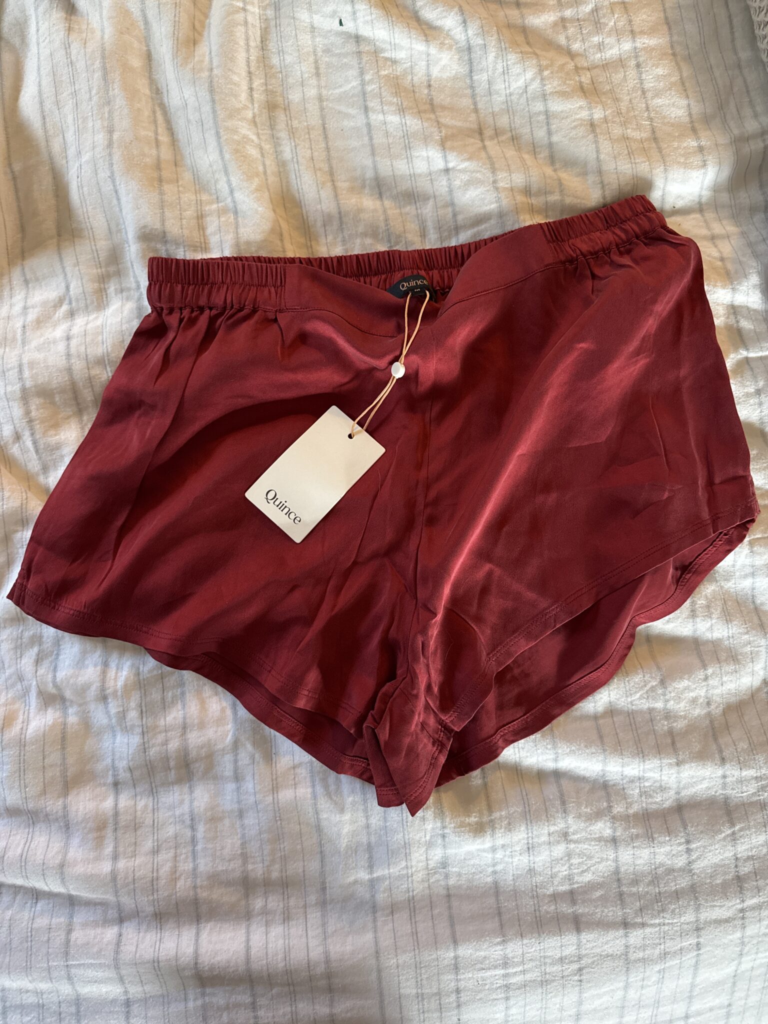 A pair of burgundy silk shorts with an elastic waistband and a white tag labeled "Cuisse," lying on a bed with a white striped sheet.