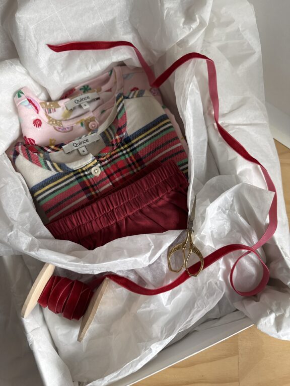 Three folded children's dresses in a gift box with tissue paper, red ribbon, and gold scissors. The dresses have different patterns and are labeled "Quince.