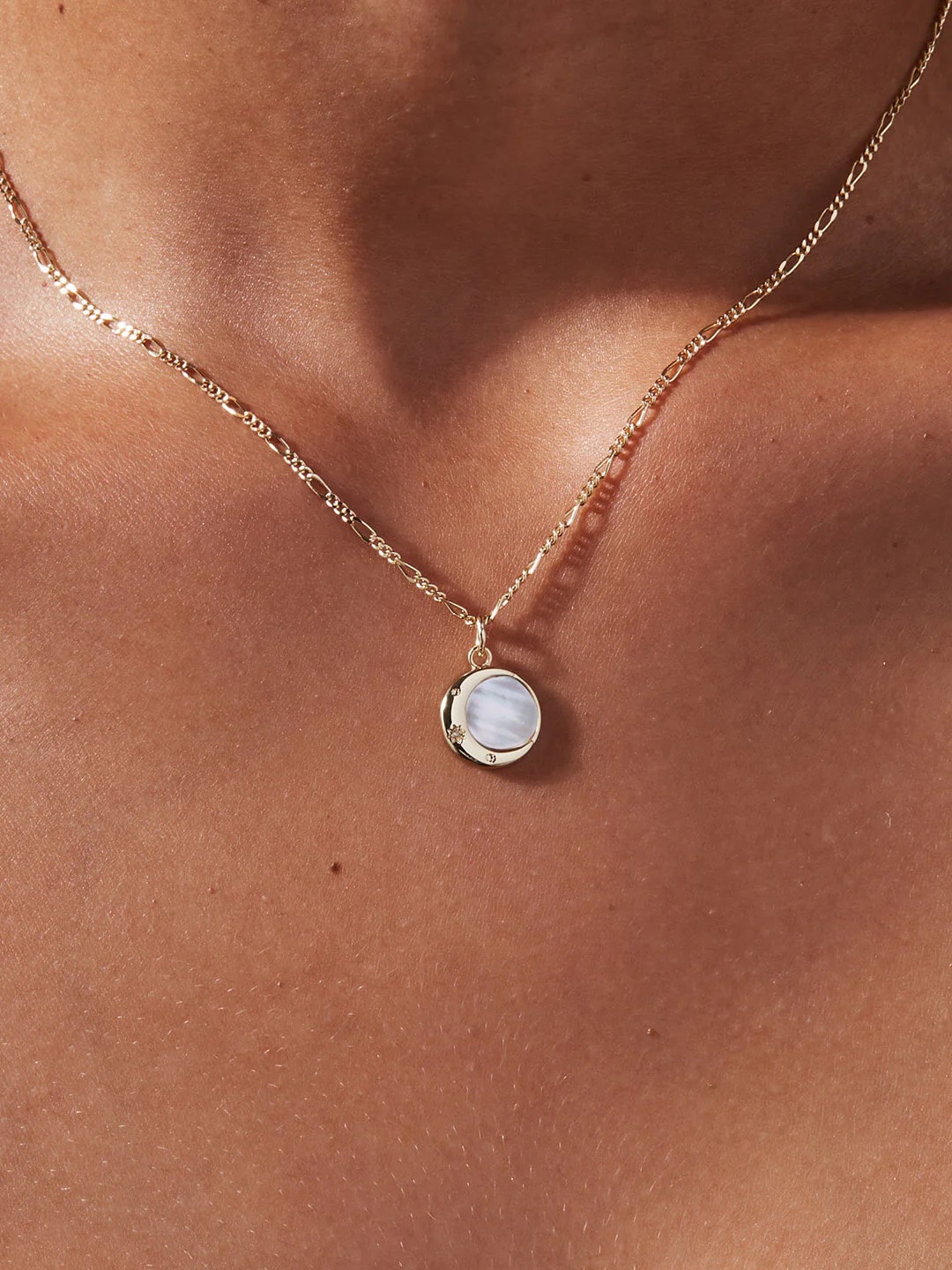 A gold chain necklace with a round, white mother-of-pearl pendant is worn around a person's neck against bare skin.