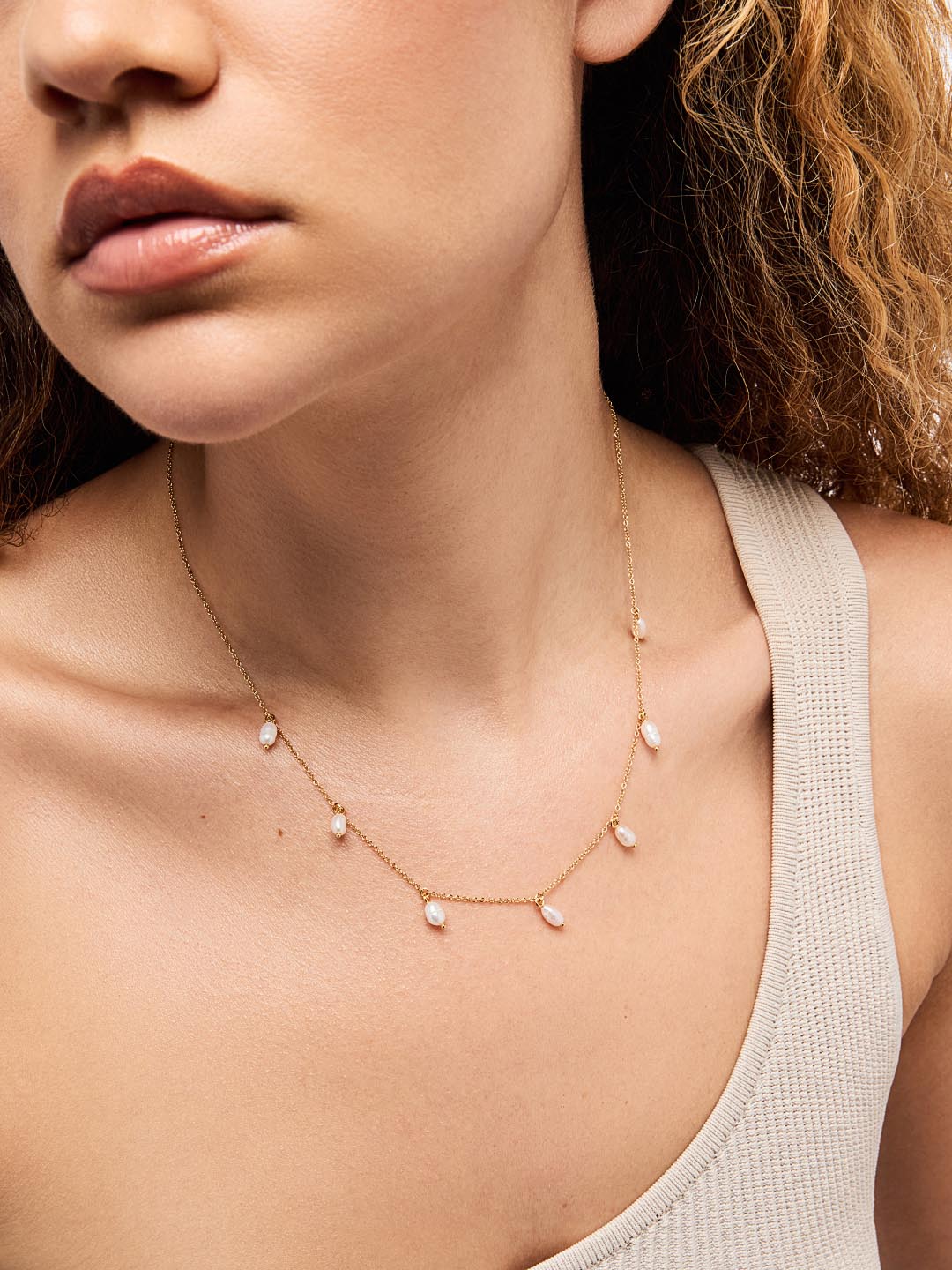 A woman wearing a one-shoulder beige top and a delicate gold necklace with small dangling pearls.
