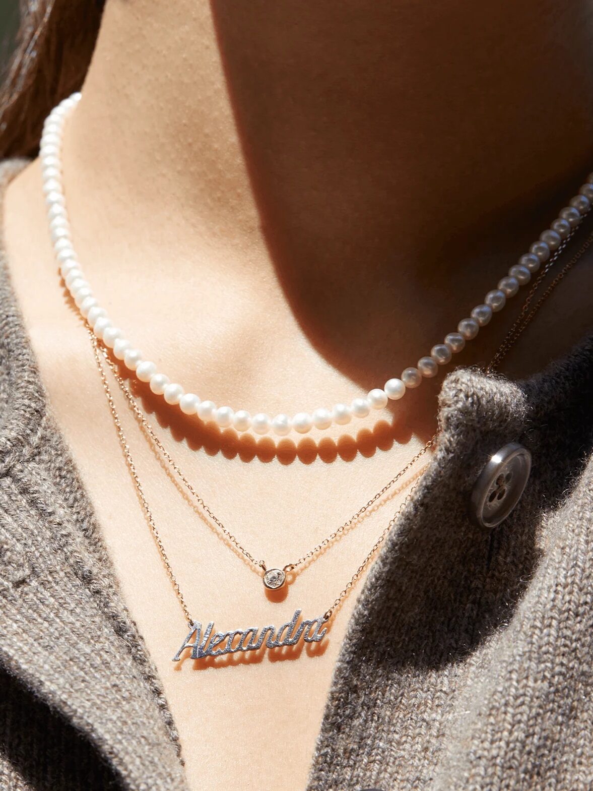 A person wearing a brown knit top with two layered gold necklaces, one with a "Alexandra" nameplate and the other with a small pendant, along with a pearl necklace.