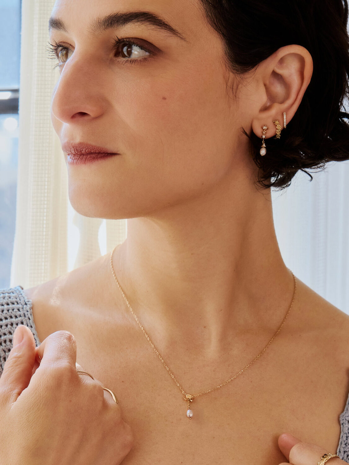 Woman wearing gold jewelry with a pearl pendant necklace and matching earrings, looking to the side in a softly lit room.