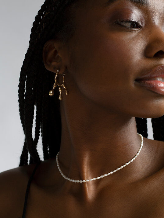 A woman with braided hair wears gold earrings, a delicate beaded necklace, and a black top, with a tattoo visible on her shoulder.