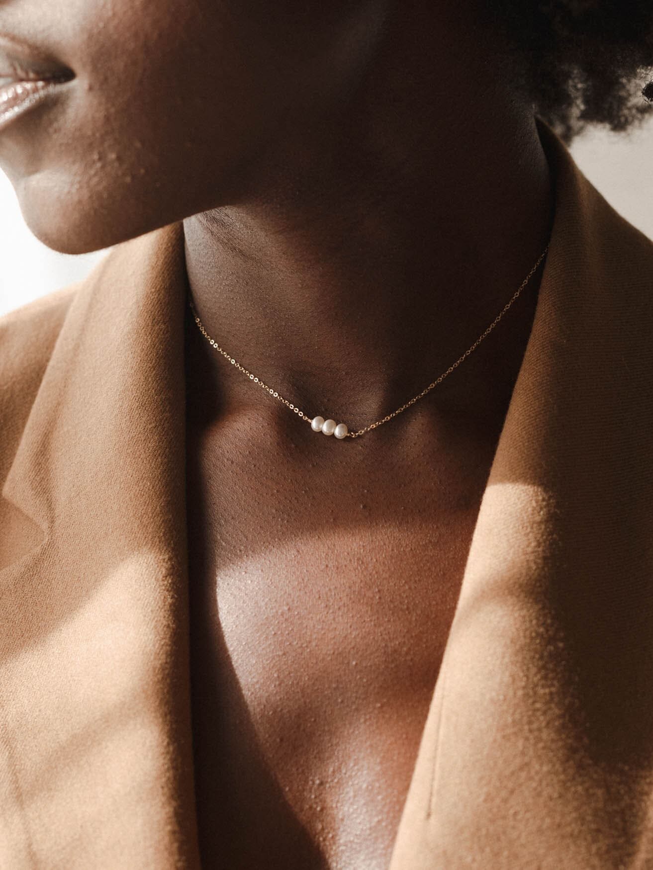 A person wearing a tan blazer and a delicate gold necklace with three small white pearls.