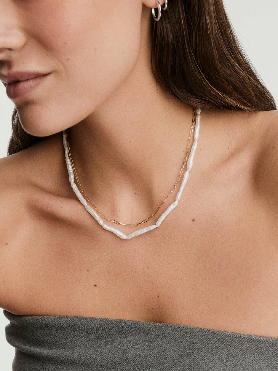 A woman wearing a strapless gray top, small hoop earring, and two layered necklaces—one with irregular white beads and one thin gold chain.