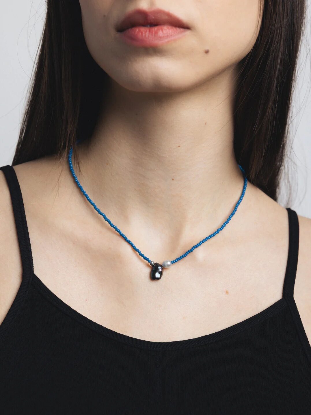 A woman wearing a black top and a blue beaded necklace with a small, dark pendant and a silver bead.