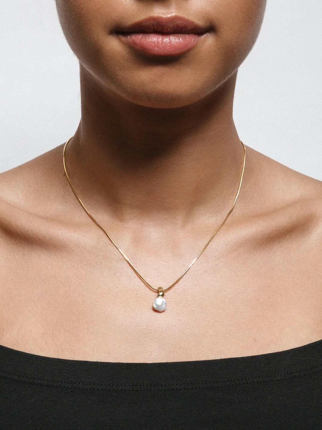 A woman wearing a thin gold necklace with a single pearl pendant, dressed in a black off-shoulder top, against a plain background.