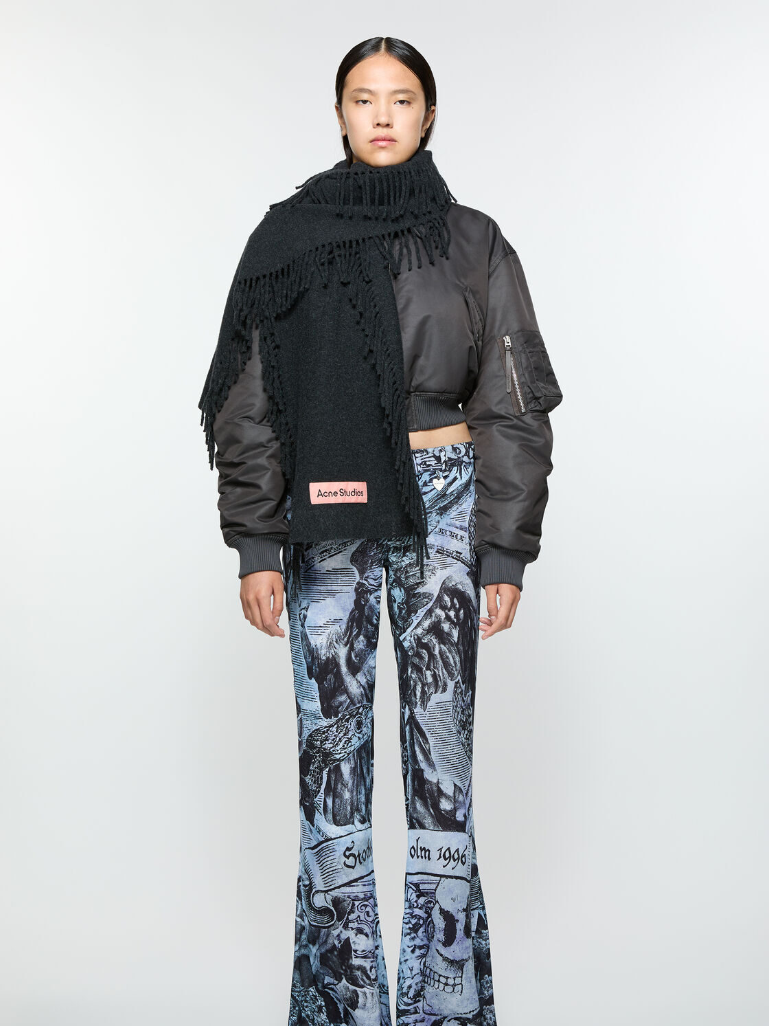 Person wearing a black bomber jacket, large black scarf, and patterned blue pants with graphics. They are standing against a plain white background.