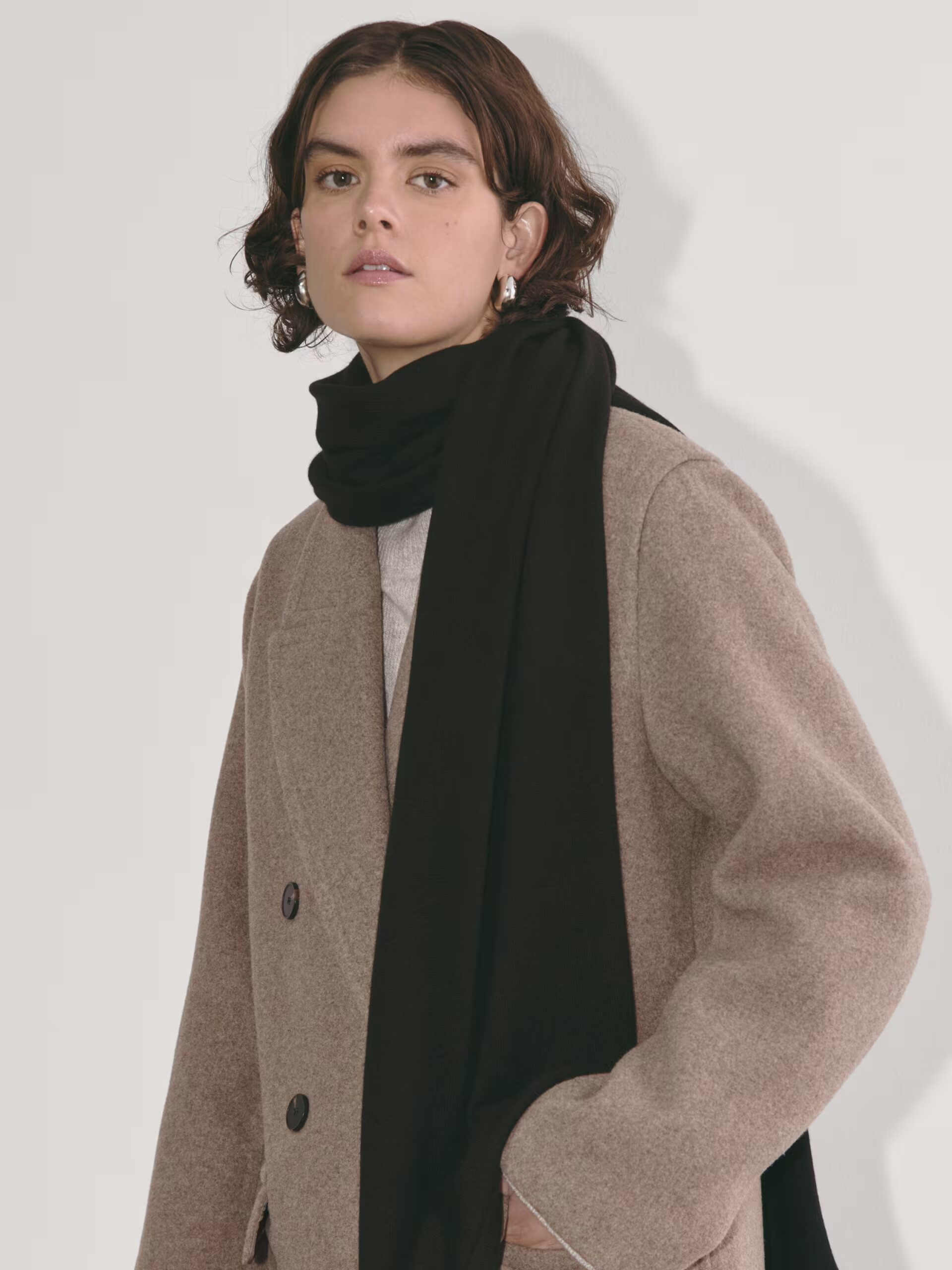 Person wearing a beige coat and a black scarf, standing against a plain background.