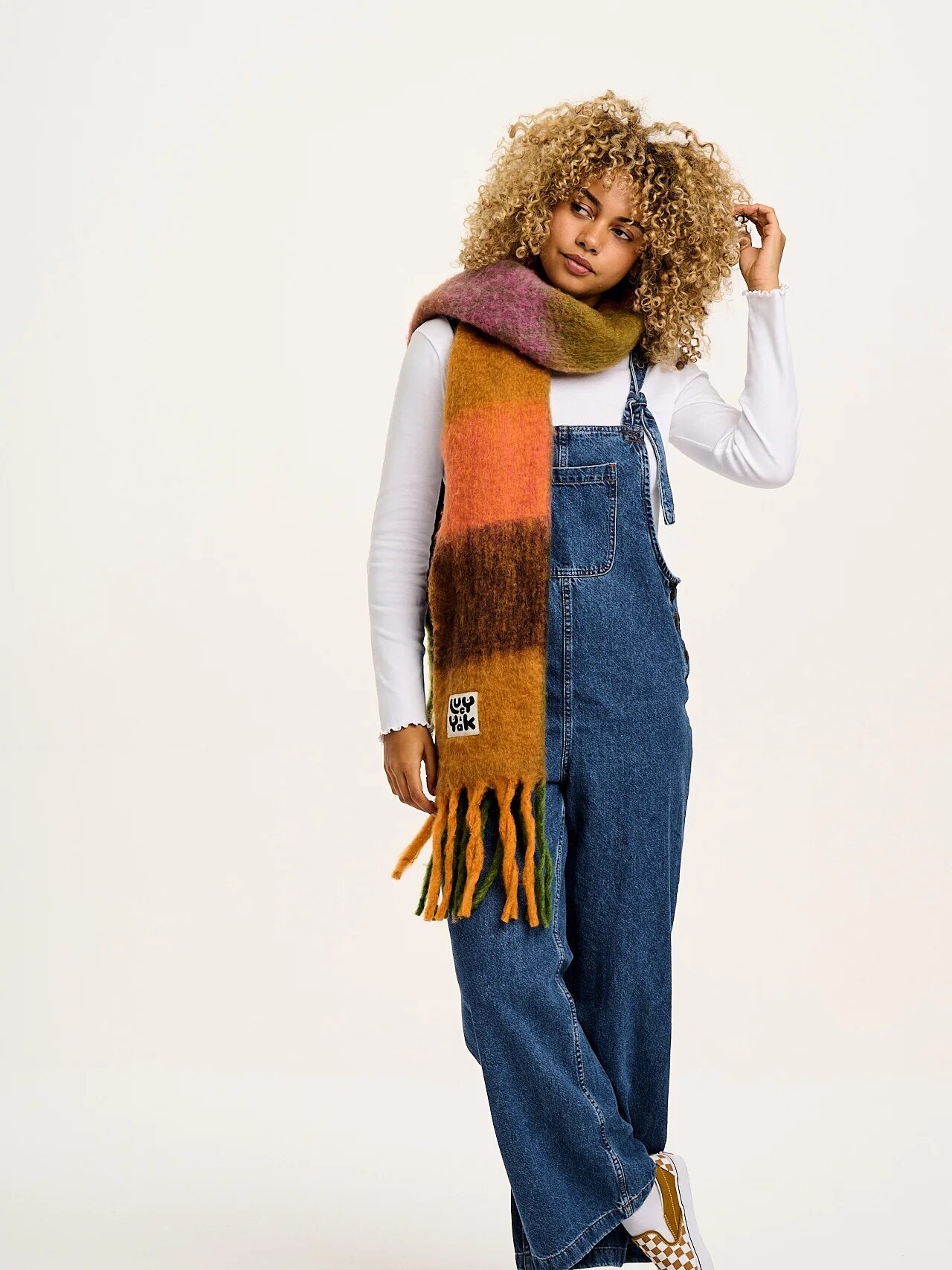 Person wearing denim overalls, a white long-sleeve shirt, a multicolored scarf, and checkered shoes, standing against a plain background.