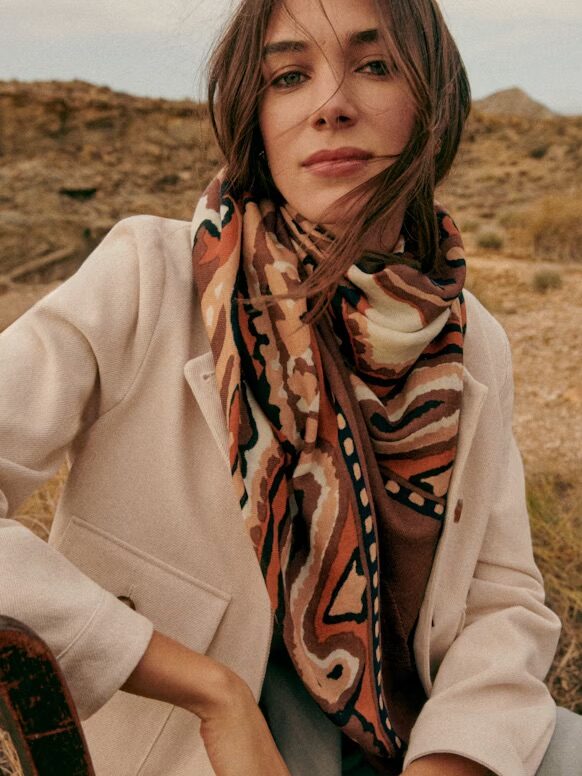 Person wearing a beige coat and patterned scarf sits outdoors in a desert landscape.