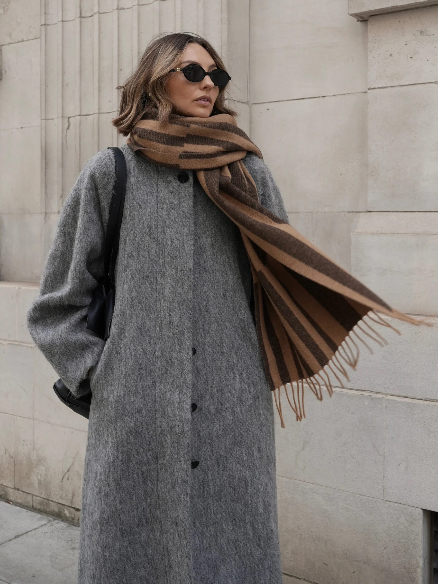 A person in sunglasses wears a long gray coat and a brown striped scarf, standing against a textured stone wall.