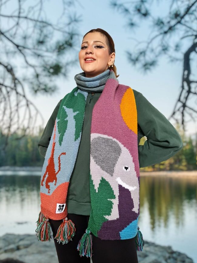 Person stands near a lake wearing a colorful scarf with animal designs, between two trees.