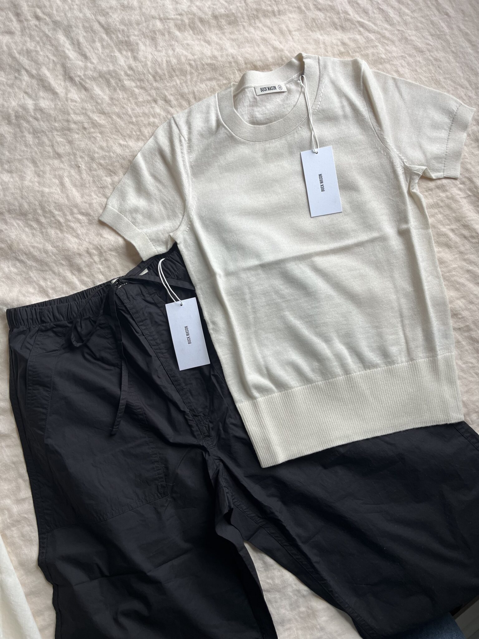 A cream short-sleeve knit shirt and black drawstring pants with tags attached, laid out on a light fabric background.