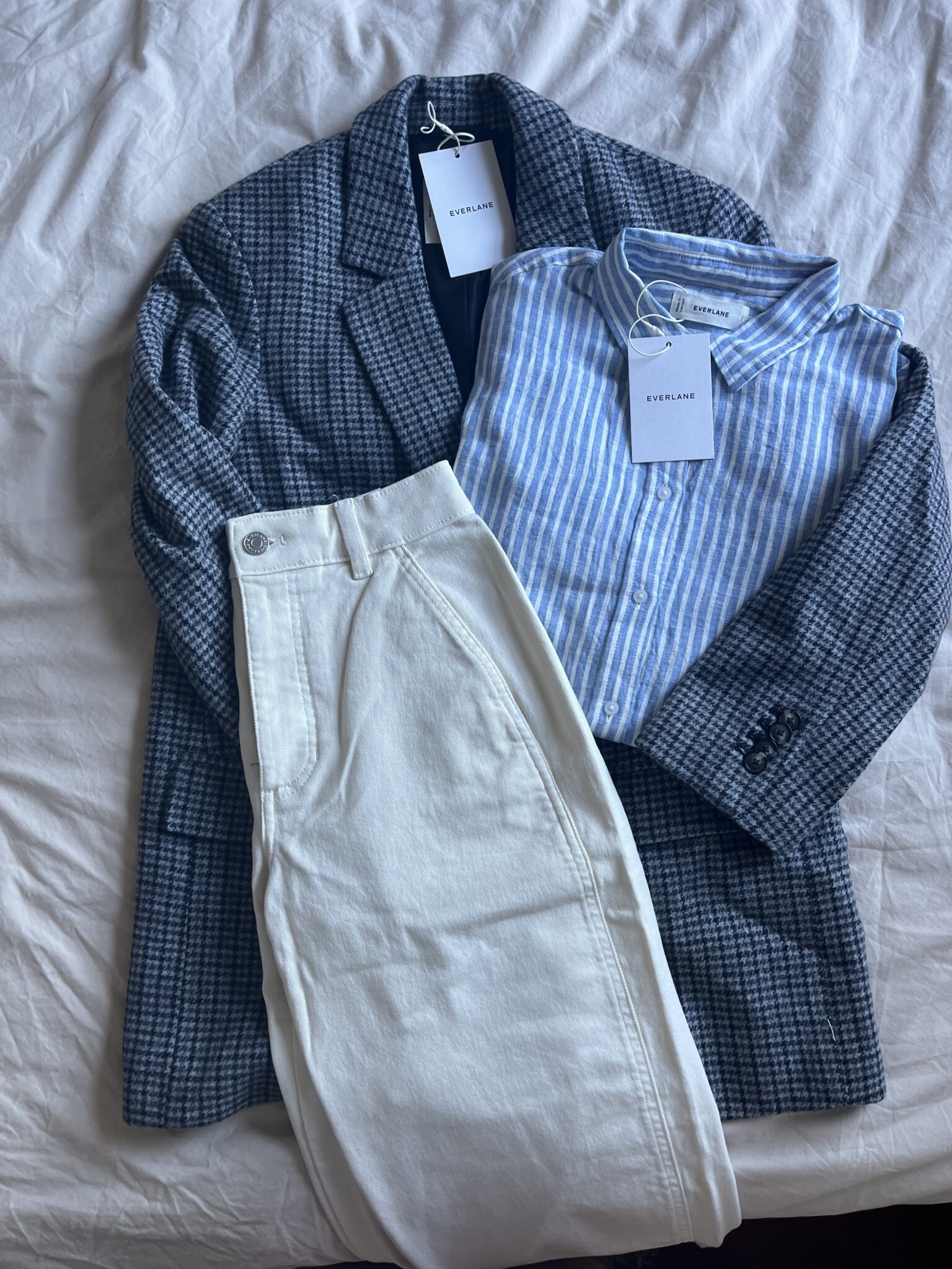 A blue checkered blazer, a blue and white striped shirt, and a pair of white pants are laid out on a bed, each with Everlane tags attached.