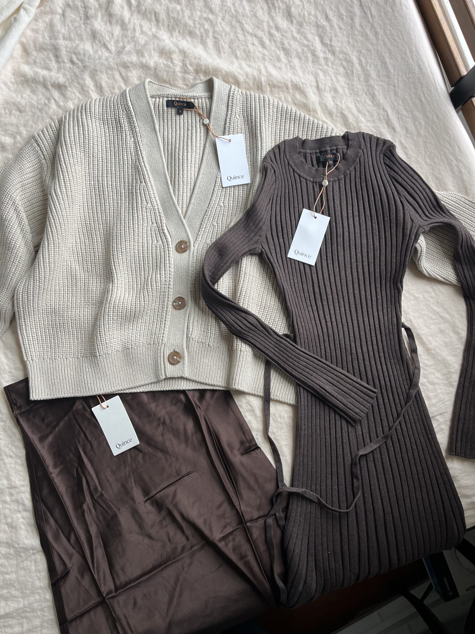 A beige button-up cardigan, a dark brown ribbed dress, and brown satin pants are laid out on a white surface, each with a tag attached.