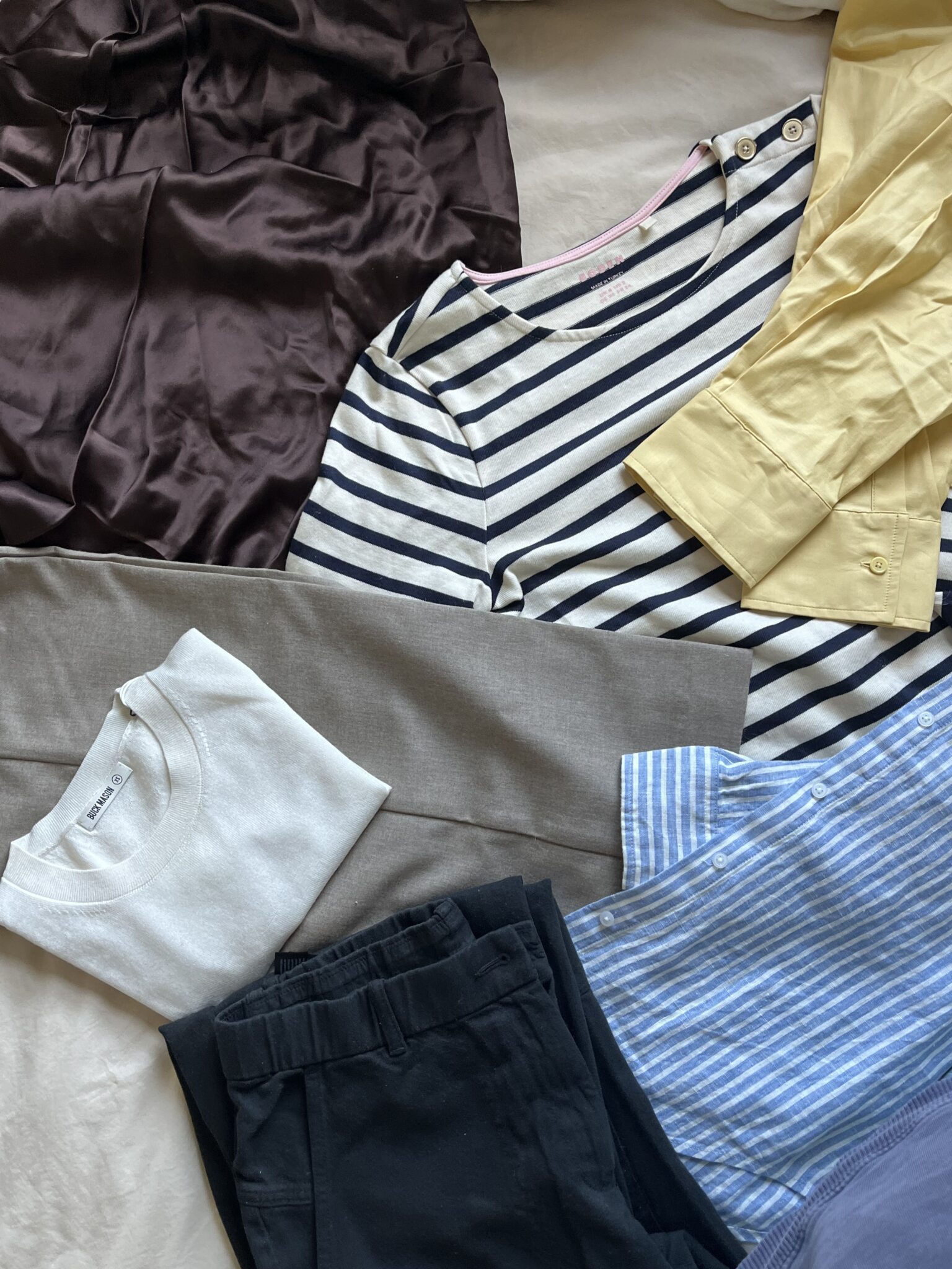 A selection of folded and laid-out clothing items, including a striped shirt, yellow blouse, blue shirt, brown skirt, gray pants, black pants, and a white t-shirt.