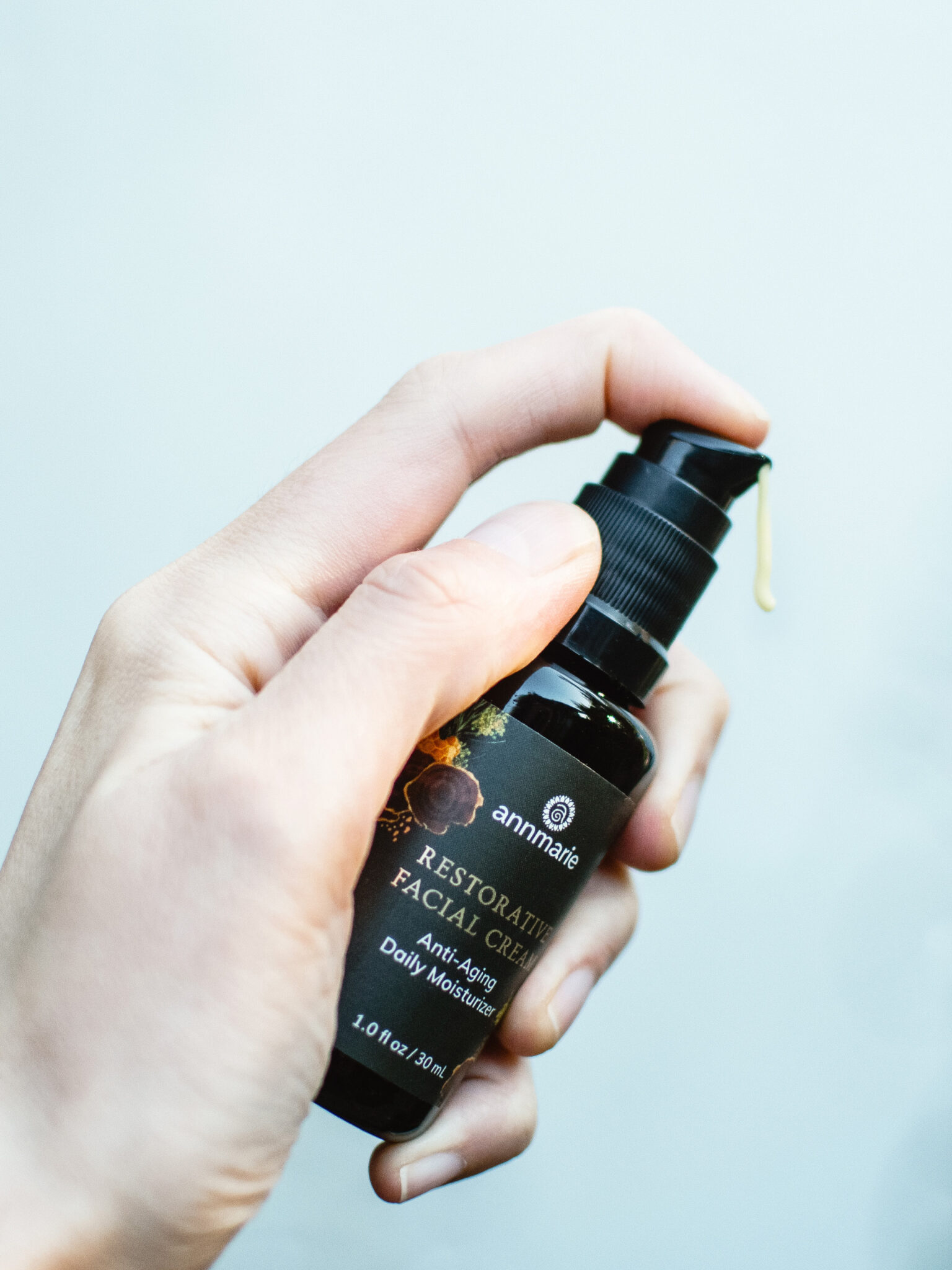 A hand holding a bottle of Annmarie Restorative Facial Cream, with a drop of cream coming out of the pump dispenser.