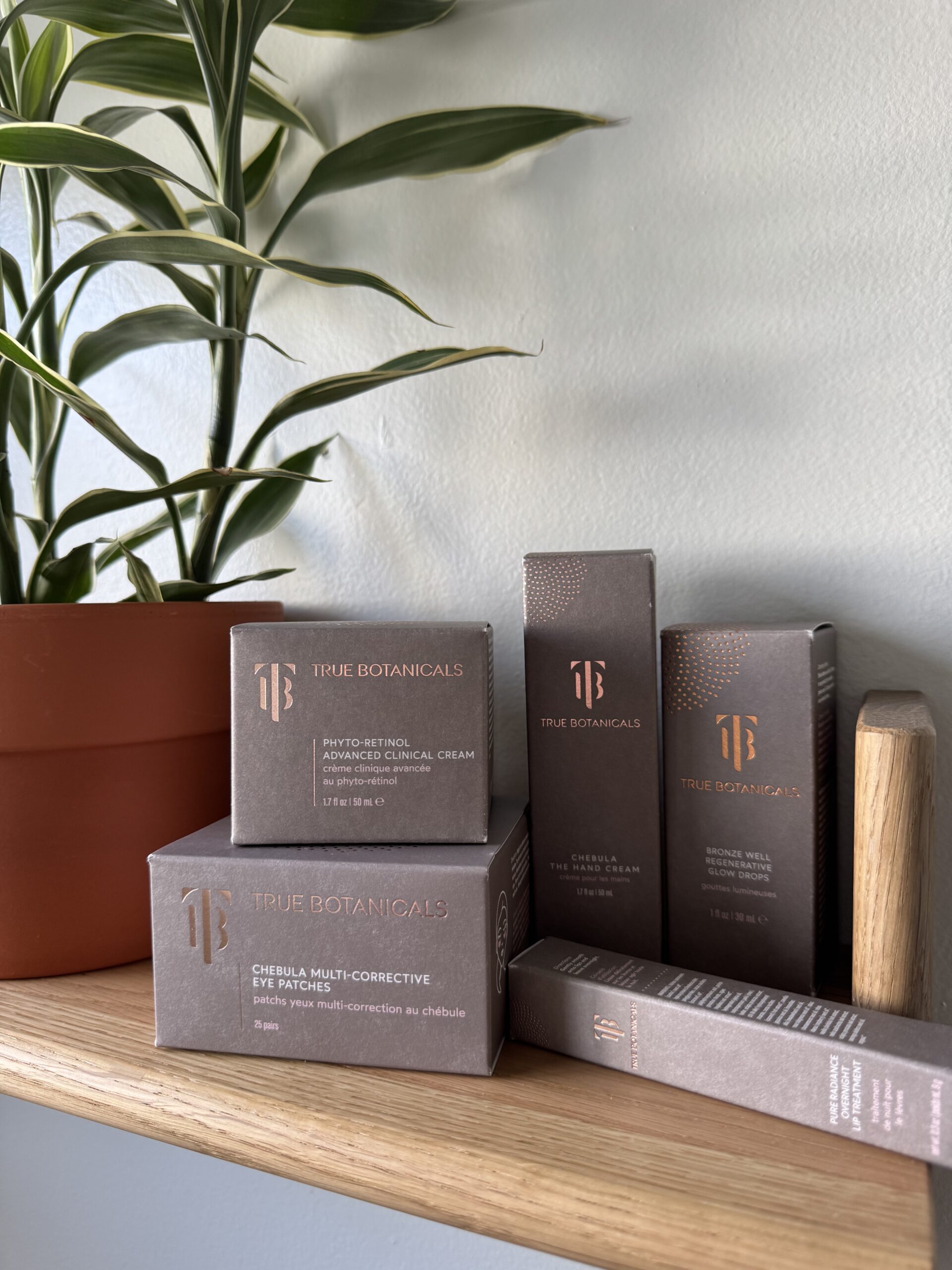 Several True Botanicals skincare product boxes are arranged on a wooden shelf next to a potted plant and leaning book, against a light-colored wall.