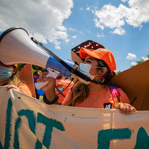 A person wearing an orange cap and face mask speaks into a megaphone while holding a banner during a daytime outdoor protest.