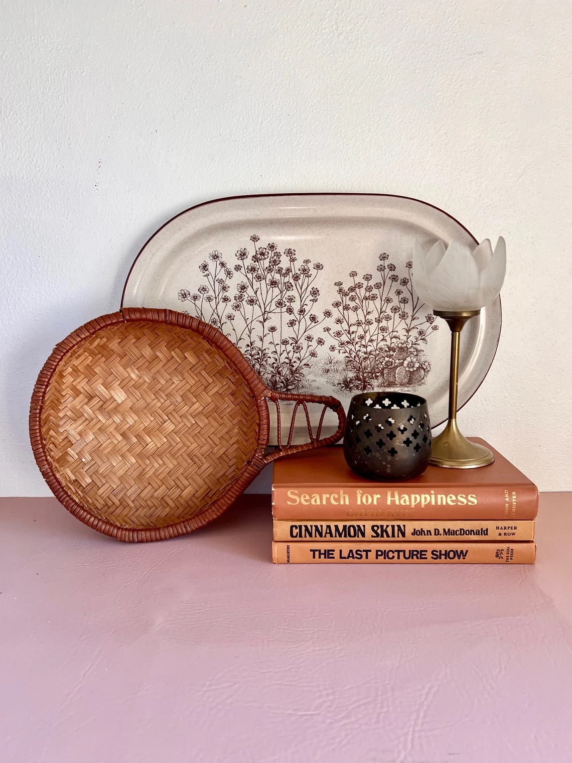 Stack of three books with a decorative plate and basket against a wall. A candle holder and a small lamp sit on top of the books.