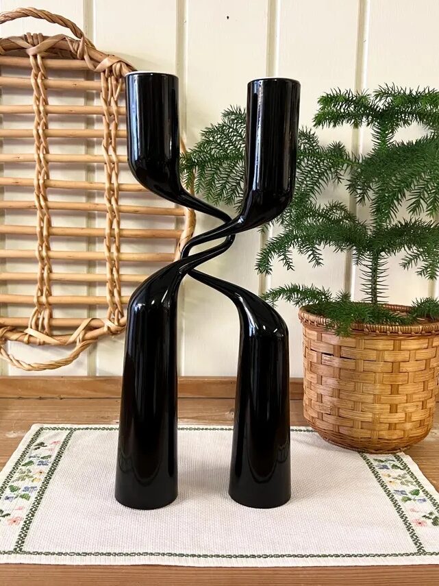 Two twisted black candle holders on a wooden table with a wicker tray and a small potted pine plant in the background.