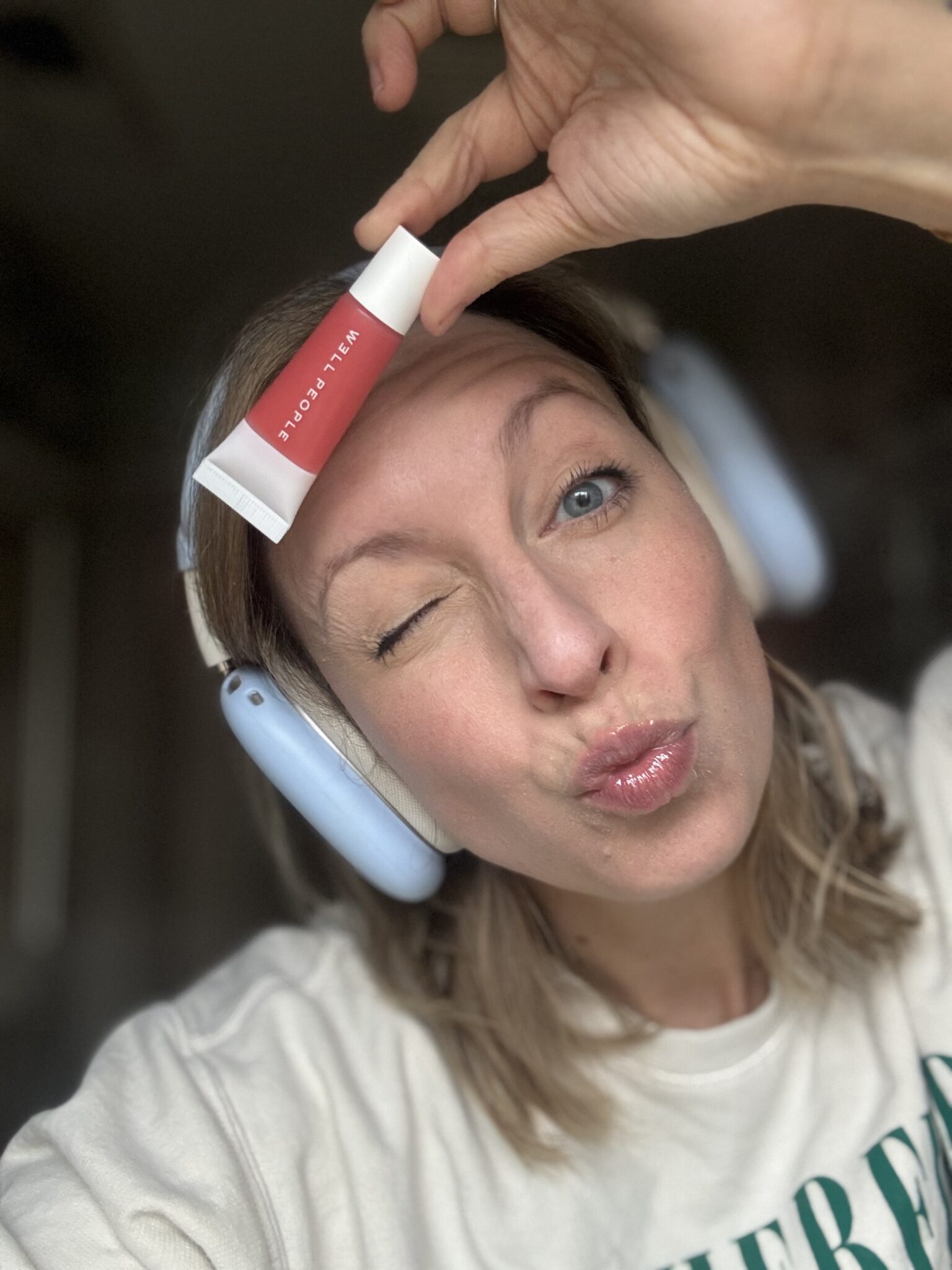 A woman wearing headphones winks and puckers her lips while holding a tube of makeup product labeled "ALL TIGERS" against her forehead.