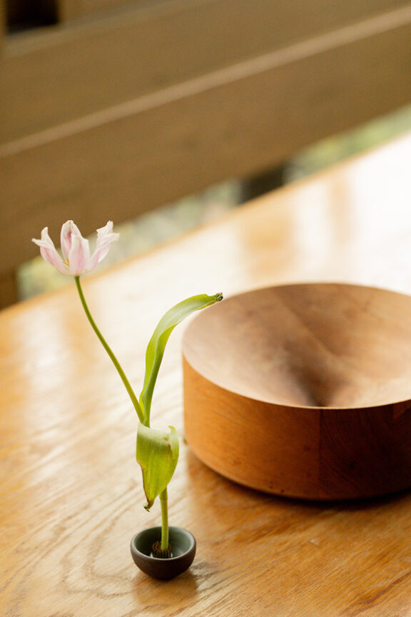 A single pink tulip with green leaves in a small vase sits on a wooden table next to a large wooden bowl.