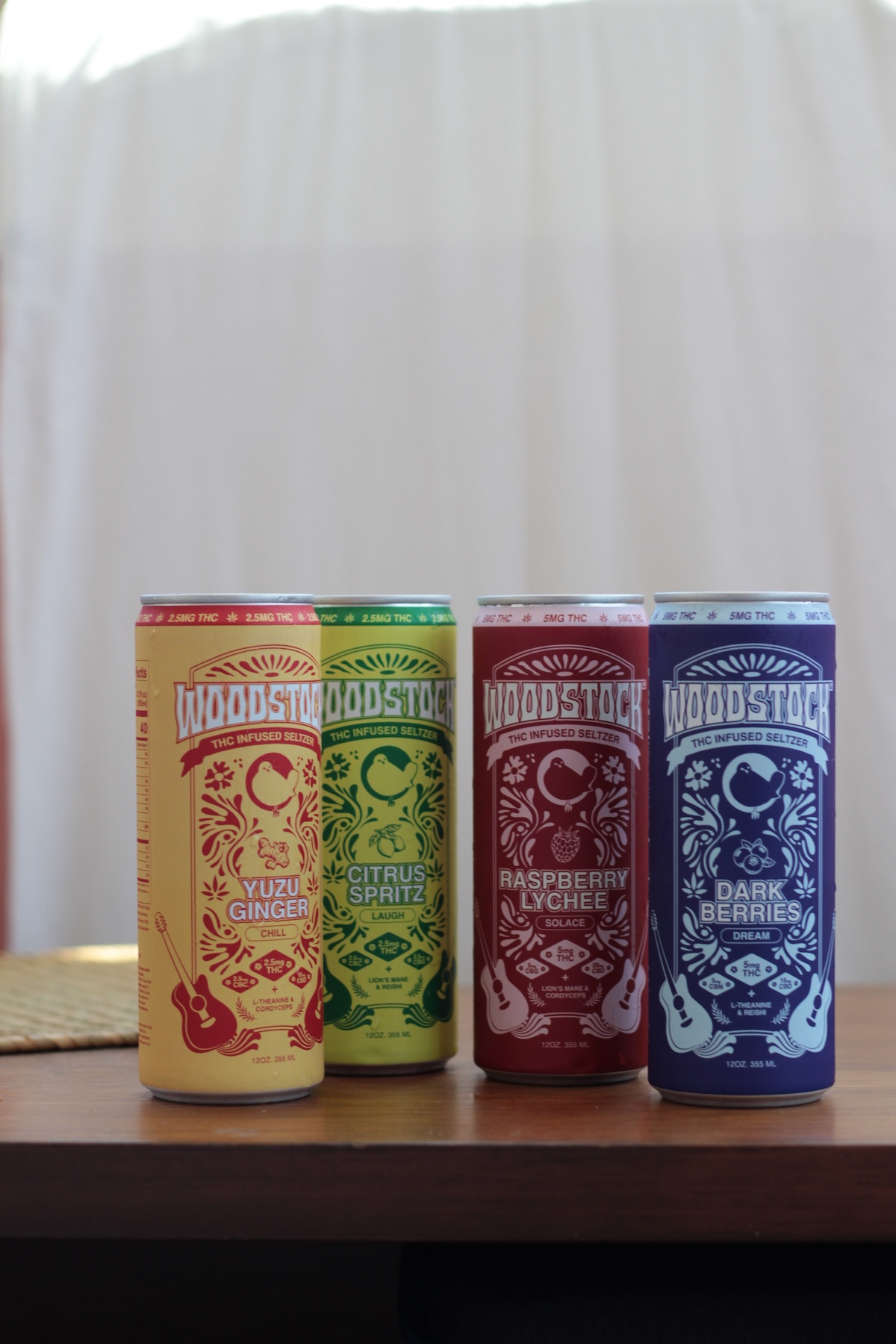 Four Woodstock beverage cans in different flavors—Yuzu Ginger, Citrus Spritz, Raspberry Lychee, and Dark Berries—are arranged in a row on a wooden surface.