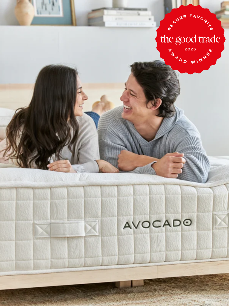 A smiling couple lies on an Avocado mattress in a bright bedroom. A red "Reader Favorite: The Good Trade 2025 Award Winner" badge is in the top right corner.