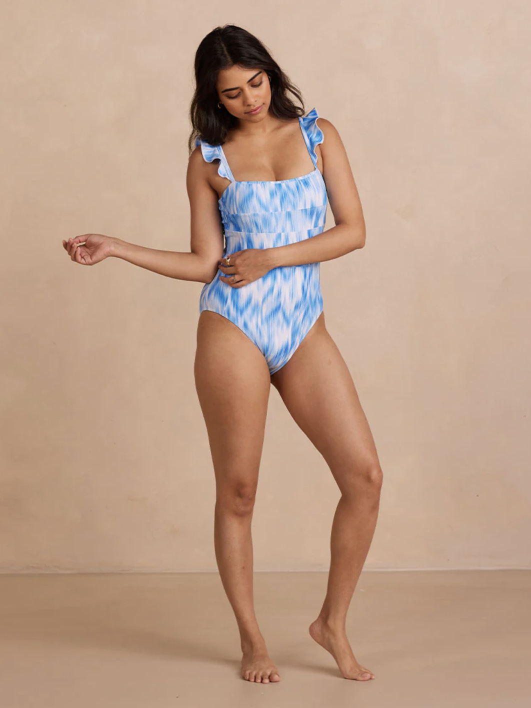 Person wearing a blue and white patterned one-piece swimsuit with ruffled straps stands against a plain beige background.