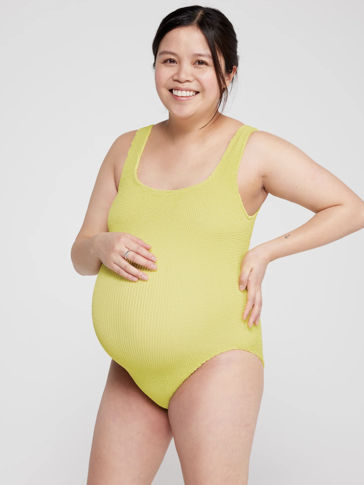 Pregnant person wearing a textured lime green swimsuit, smiling and resting one hand on their hip and the other on their belly, against a plain background.
