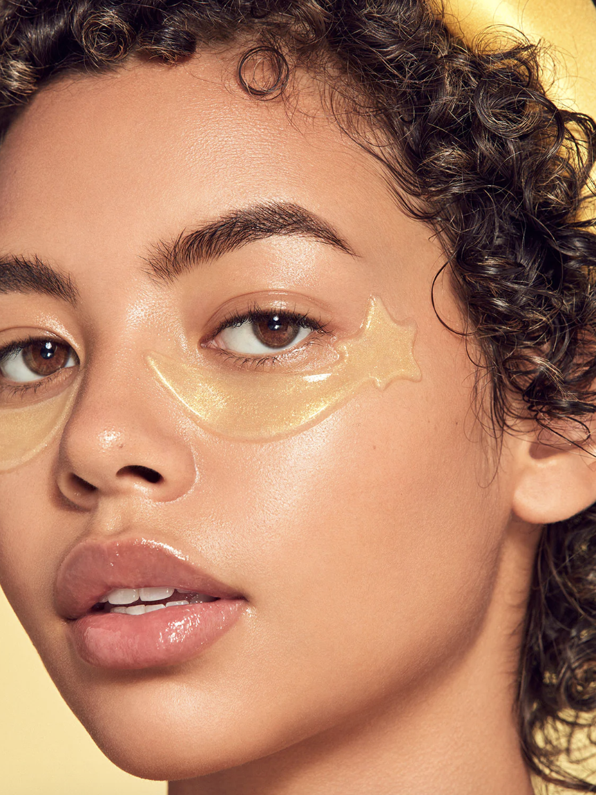 A person with curly hair wears gold under-eye gel patches shaped like shooting stars against a yellow background.