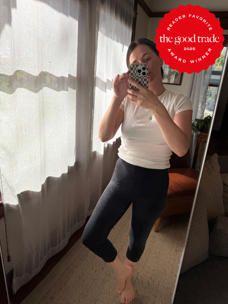 Person in a white top and black leggings takes a mirror selfie in a room with sheer curtains; a red "The Good Trade Award Winner 2025" badge appears in the top right corner.
