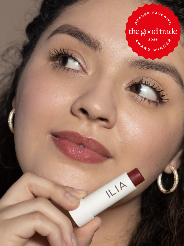 Person holding a tube of ILIA lip product, looking upward. The image features a "Reader Favorite the good trade 2025 Award Winner" badge in the corner.