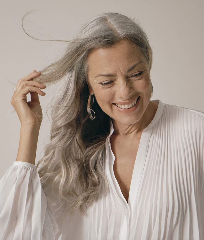 A woman with long gray hair smiles, wearing a white pleated blouse, against a plain background.