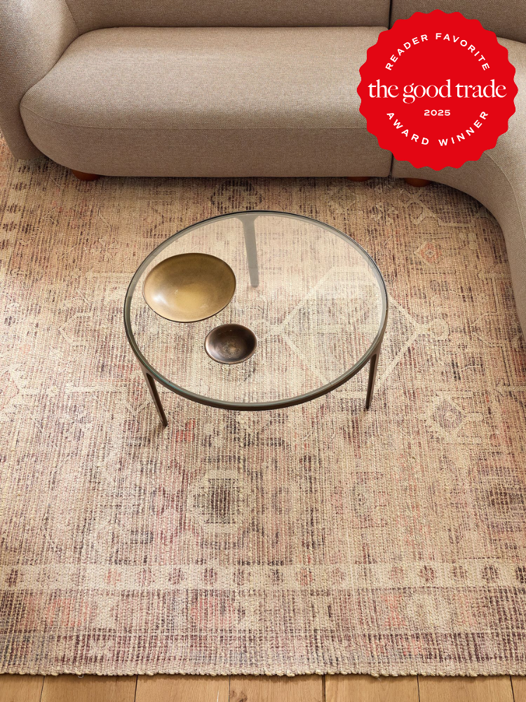 A round glass coffee table with two brass dishes sits on a patterned rug in front of a beige sofa. A "The Good Trade 2025 Award Winner" badge is in the upper right corner.