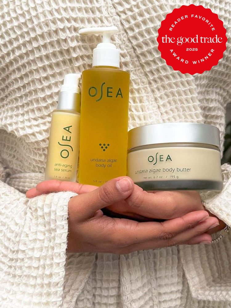 Person in a white textured robe holds OSEA skincare products: anti-aging sea serum, body oil, and body butter. A red "The Good Trade 2025 Reader Favorite Award Winner" badge is in the corner.