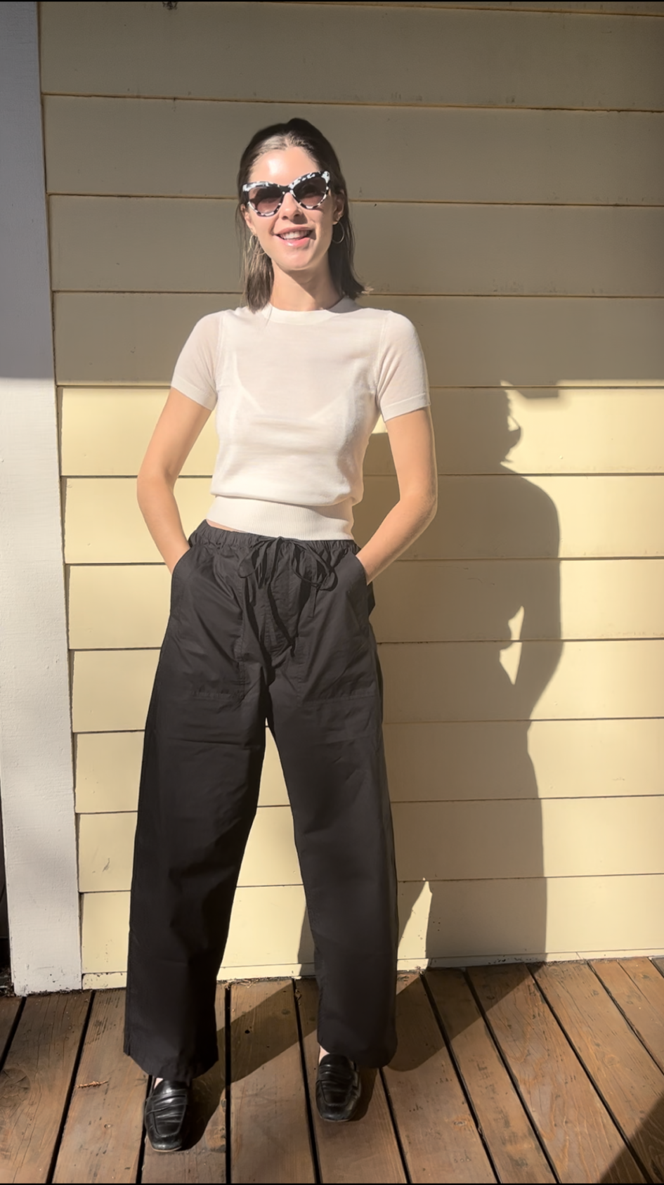 A person stands on a wooden deck against a yellow wall, wearing a white t-shirt, black pants, black shoes, and white sunglasses, with hands in pockets and sunlight casting a shadow.