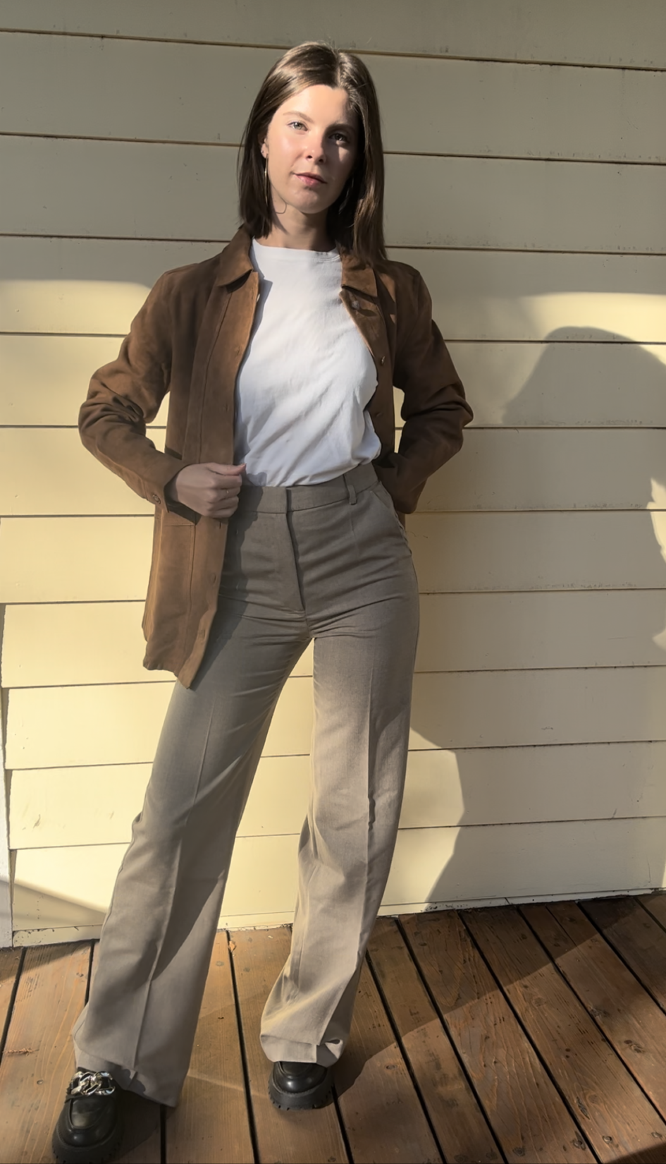 A woman stands on a wooden deck against a light-colored wall, wearing a white shirt, brown jacket, beige pants, and black shoes. She looks slightly to the side.