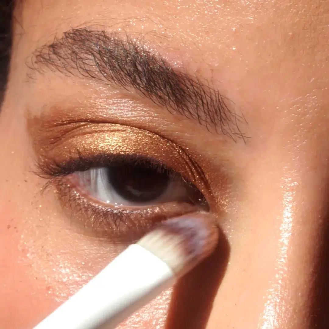 A close-up of a person applying shimmery bronze eyeshadow to their eyelid with a makeup brush.