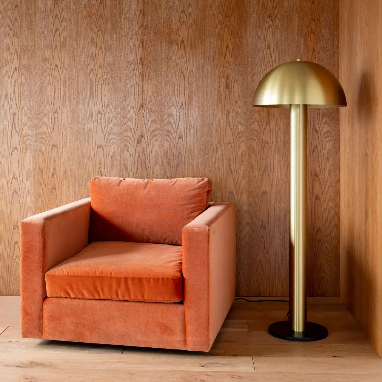 An orange velvet armchair sits next to a tall brass floor lamp with a dome-shaped shade against a wood-paneled wall.