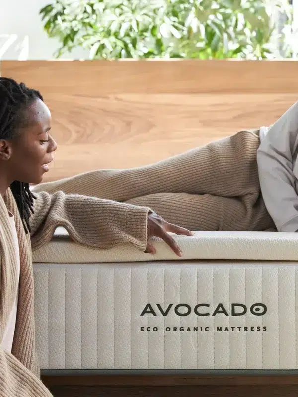 Two women sit and recline on a bed with an Avocado Eco Organic Mattress, with plants visible in the background.