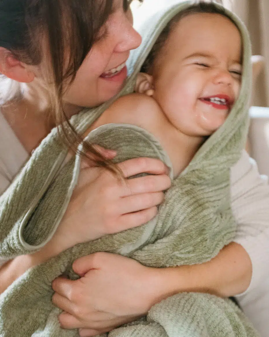 Adult holding a smiling toddler wrapped in a green towel, both appearing happy and engaged in a light moment together indoors.