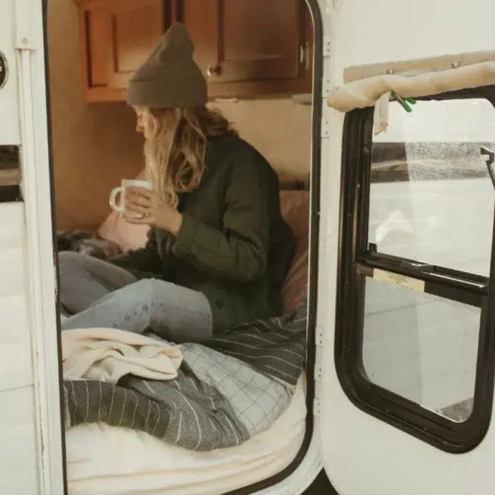 A person wearing a jacket and beanie sits on a bed inside a camper, holding a mug and looking out the open door.