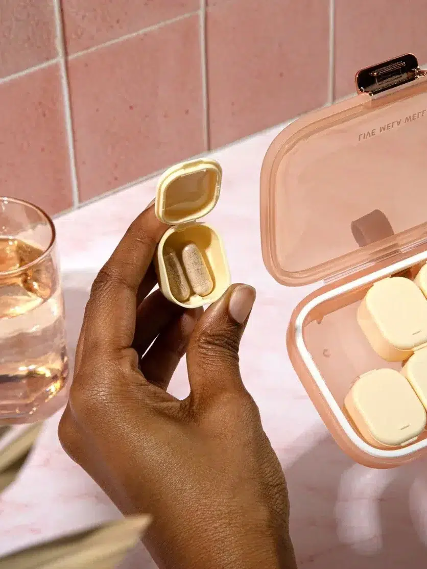 Hands holding a pill case next to a glass of water on a pink countertop with a larger pill organizer.