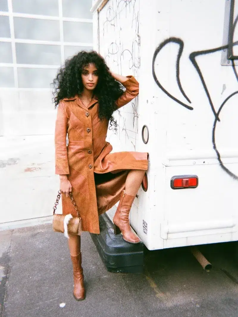 Woman in a brown coat and boots leans against a graffiti-covered white truck, holding a purse.