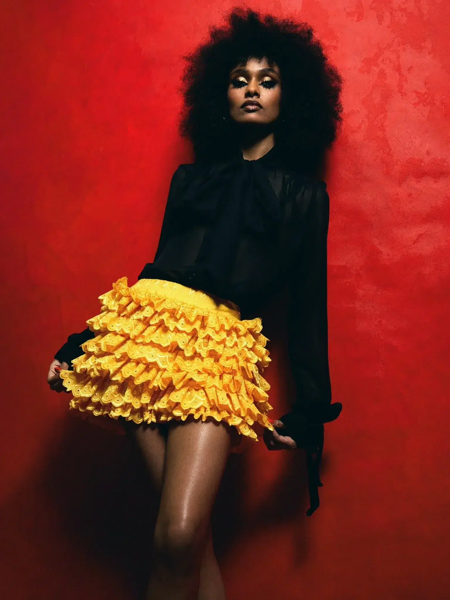 Person with curly hair wearing a black blouse and a yellow ruffled skirt stands against a red textured background.